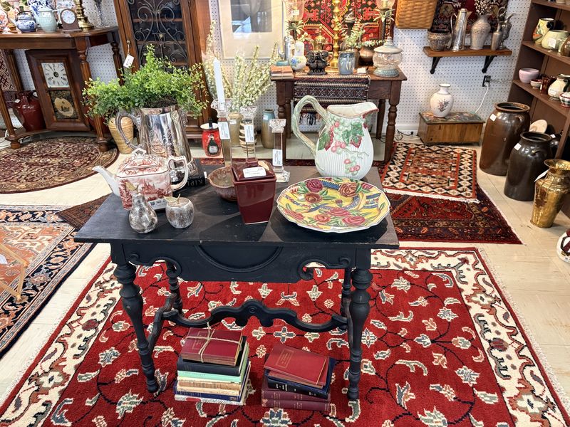A black table set with pottery, glass, and a stack of old books on a red Oriental rug, with more rugs and antiques behind it